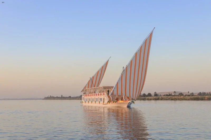 A luxury traditional Egyptian Dahabiya wooden sailing boat cruising along the lush palm-lined banks of the Nile River.