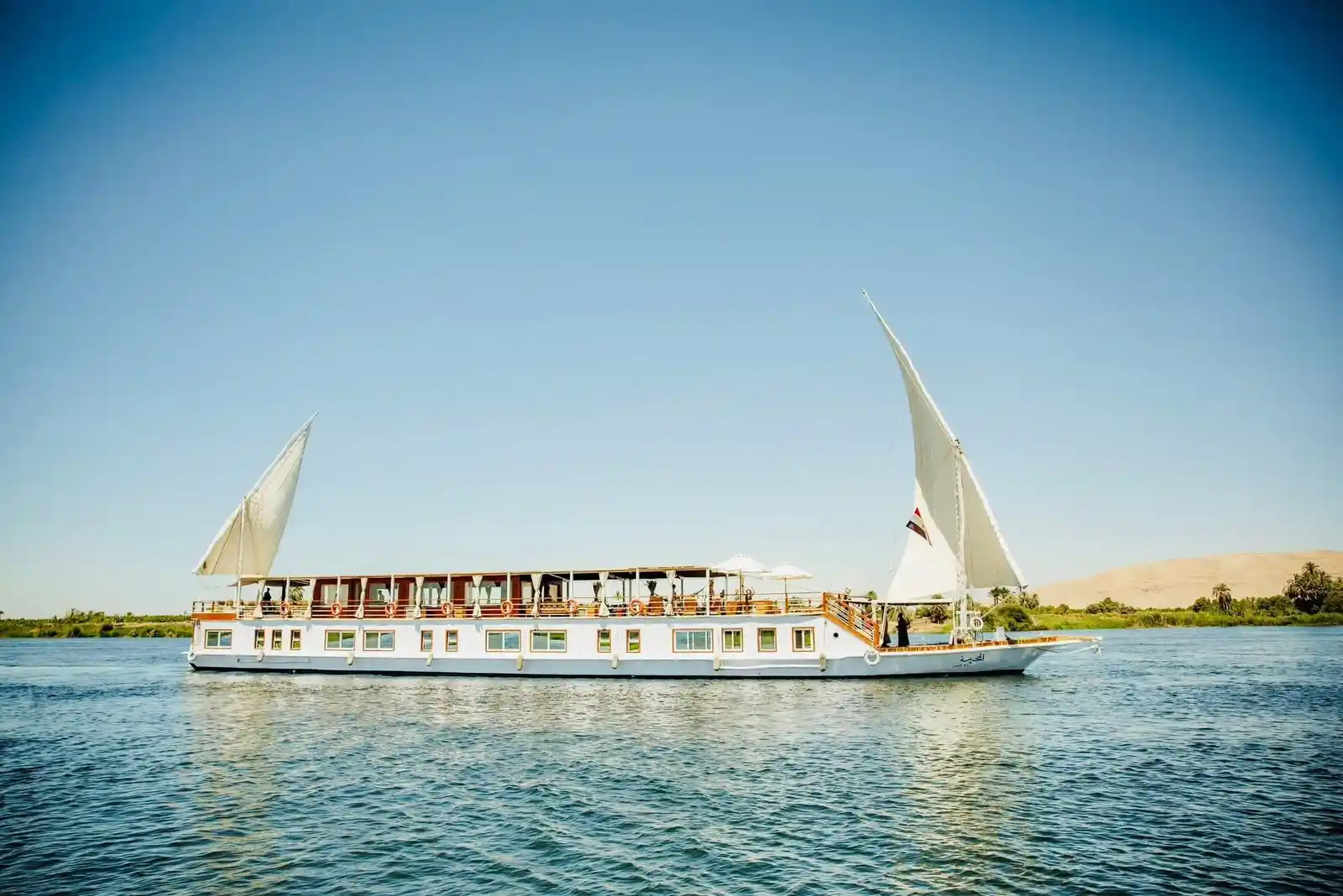 A classic luxury Dahabiya sailing boat on a premium Nile cruise tour passing scenic historic riverbanks in Egypt.