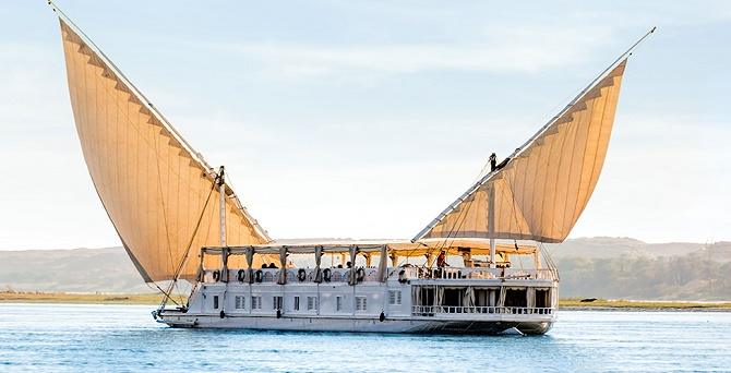 A traditional Dahabiya sailing on the Nile River, surrounded by desert cliffs and lush riverbanks.
