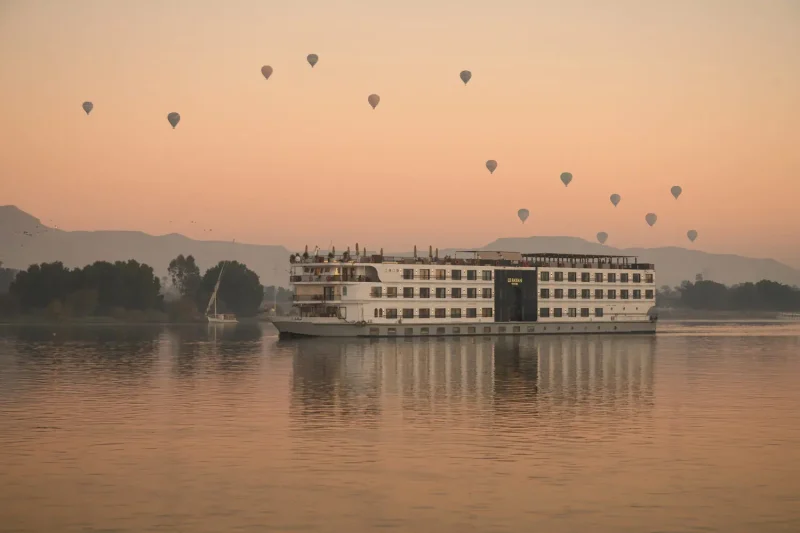 Classic white Nile cruise ship sailing on the calm blue waters of the Nile River in Egypt during a vibrant sunrise with hot air balloons floating in the clear sky.