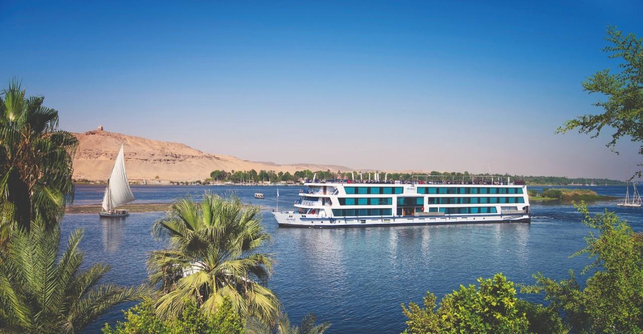 Luxury cruise ship sailing on the Nile River at sunset with palm trees in the background.