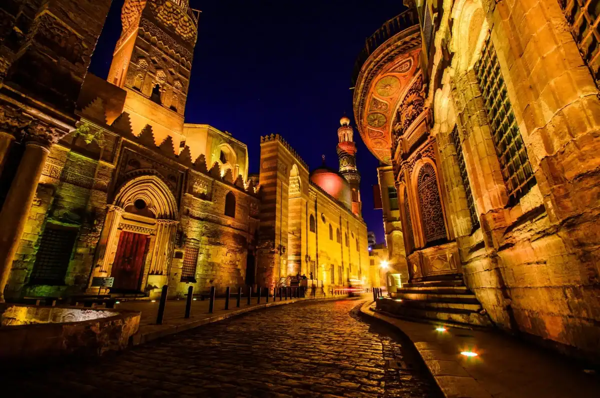 A panoramic view of El Moez Street in Islamic Cairo, Egypt, showing the historic Al-Muizz architecture and the Qalawun Complex under a blue sky.