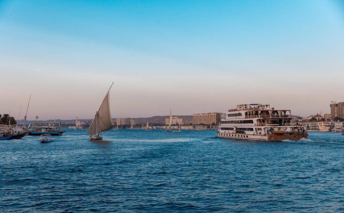 A wide scenic view of the Nile River in Egypt featuring a traditional wooden Felucca boat sailing past lush green banks and ancient desert hills at sunset.