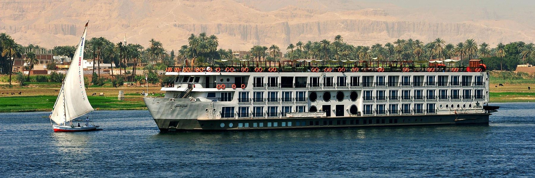 A luxury Nile cruise ship sailing on the river in Egypt past palm trees and ancient temples at sunset.