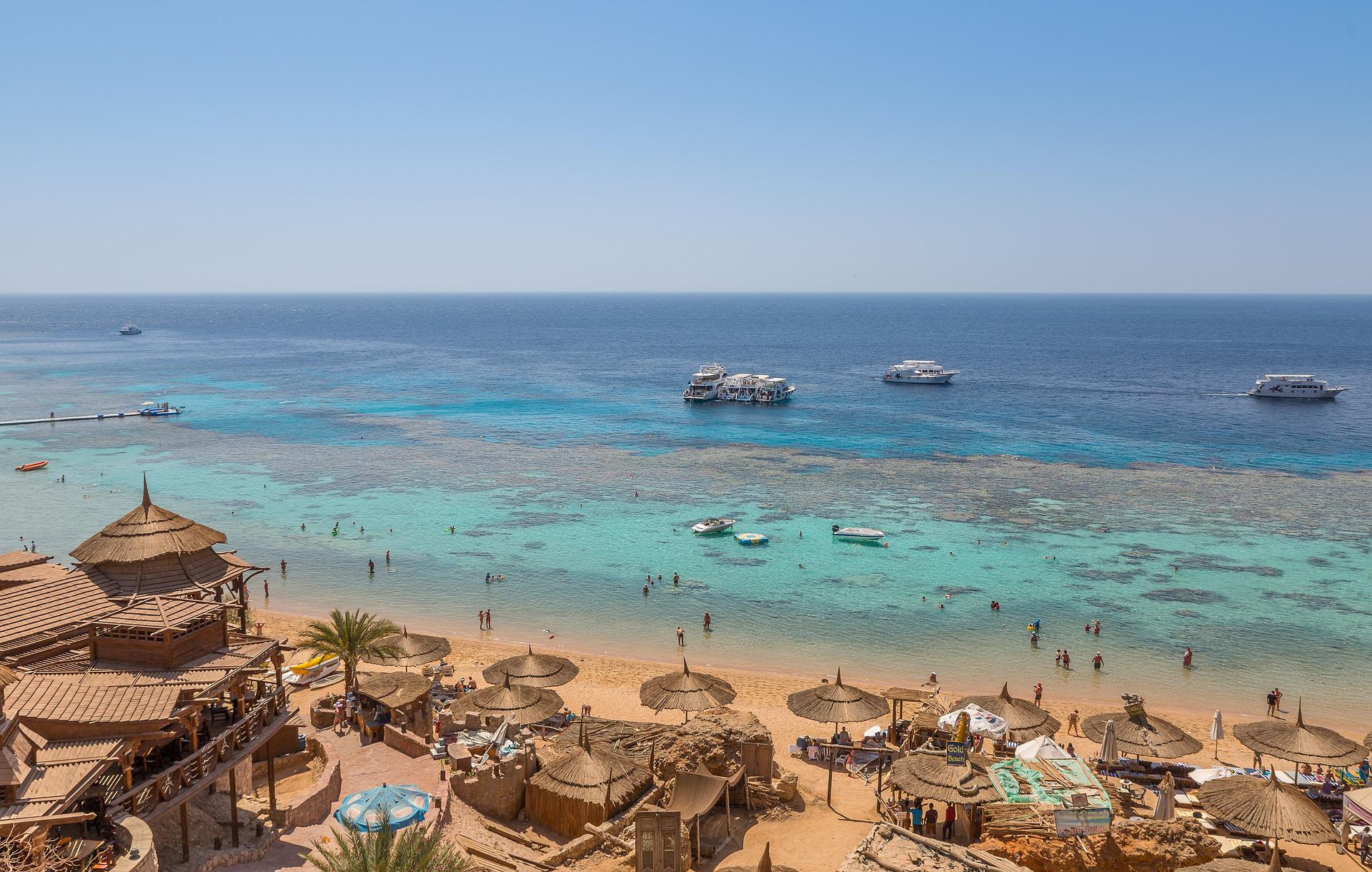 A panoramic, scenic view of Sharm El Sheikh showing the crystal-clear turquoise waters of the Red Sea, luxury coastal resorts, and the rugged desert mountains of the Sinai Peninsula in the background.
