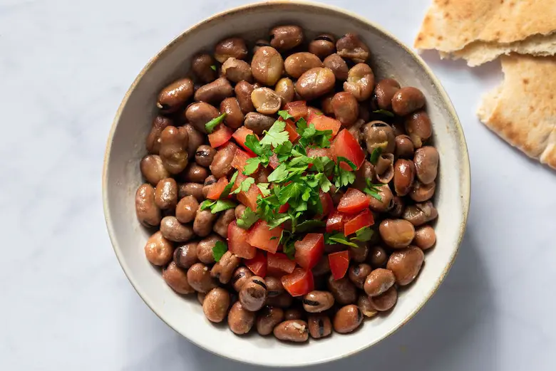 A bowl of cooked fava beans topped with chopped diced tomatoes and fresh parsley, served next to warm pita bread.