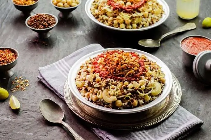 A bowl of traditional Egyptian Koshari layered with rice, lentils, macaroni, chickpeas, red tomato sauce, and crispy fried onions, surrounded by spice dishes and lemon wedges.
