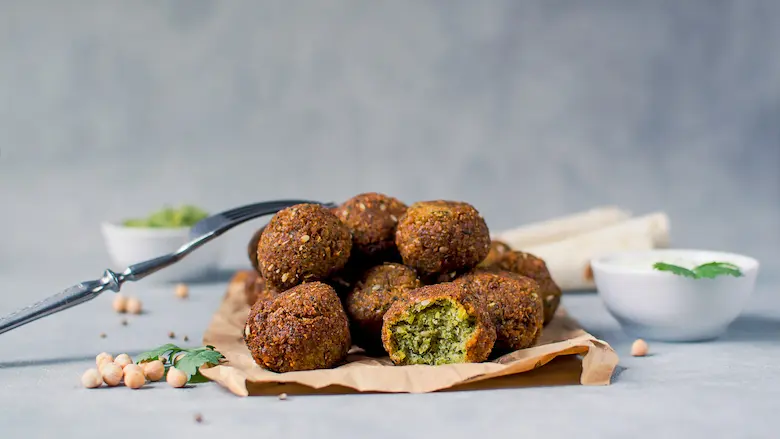 A stack of crispy fried Egyptian Ta'ameya falafel balls on parchment paper, showing one broken open to reveal its bright green herb interior.